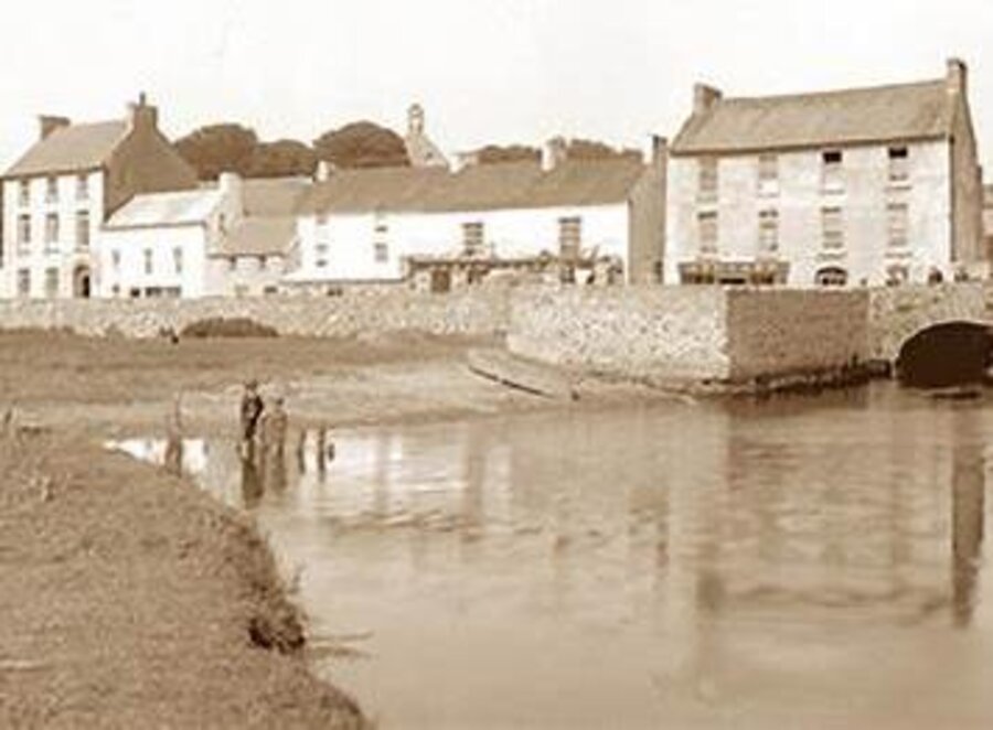 A sepia photograph of Castlebridge village around 1930, with the buildings reflected in the river